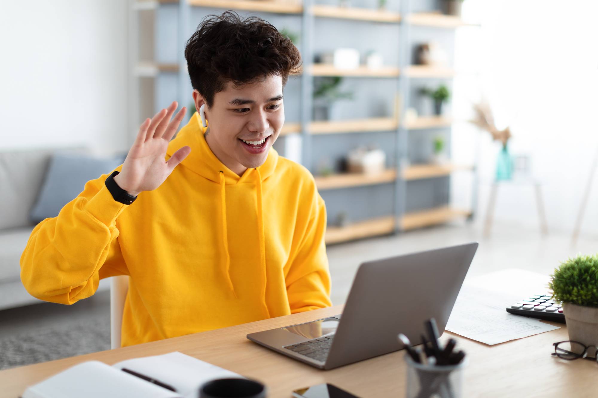 Portrait of happy asian male in bright yellow hoodie making having video call sitting at desk at home office and talking with colleagues or family, waving hand to webcam.