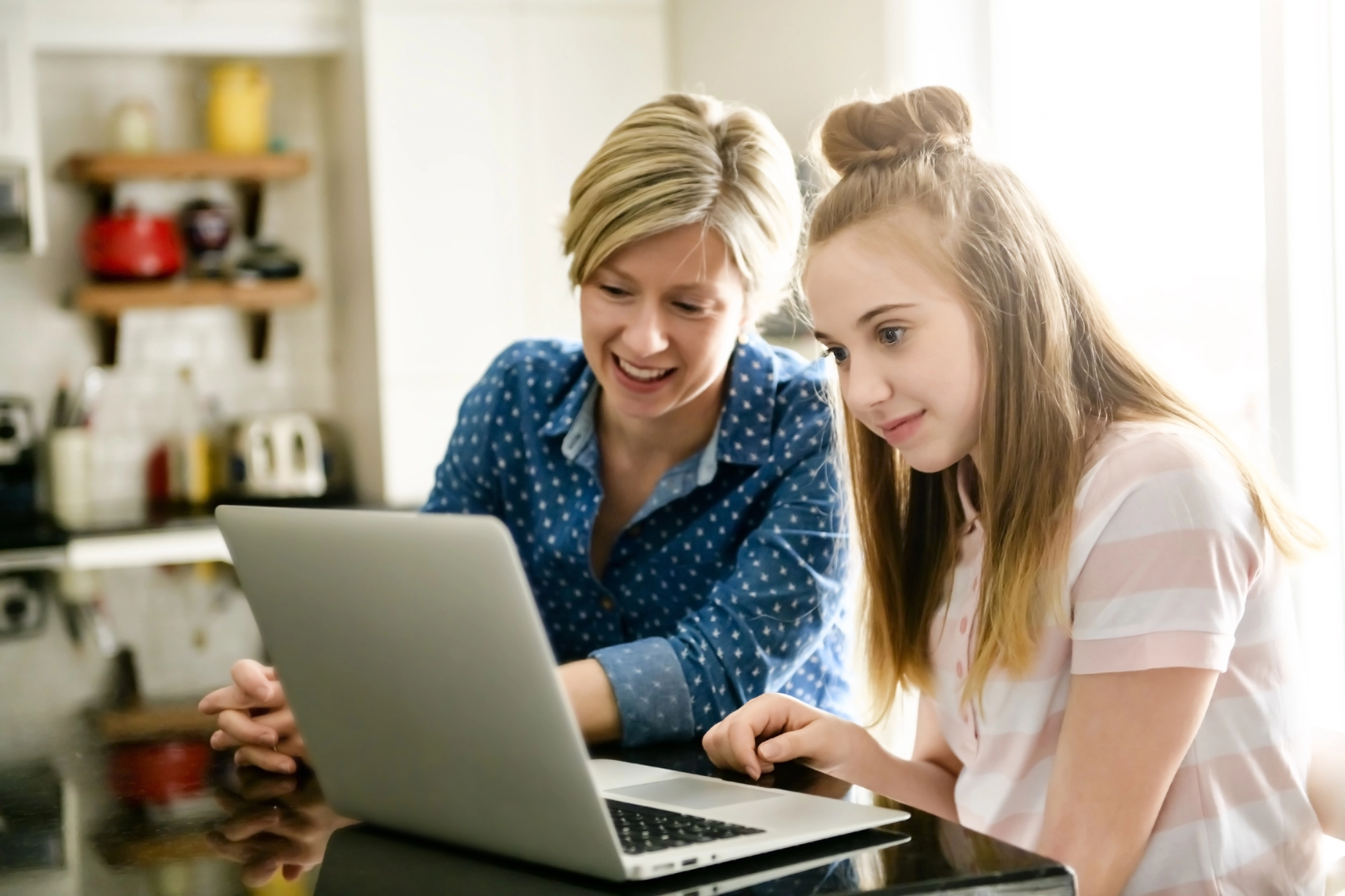 Mother and teenage daughter smiling while looking at a laptop together, planning senior year college applications at home.