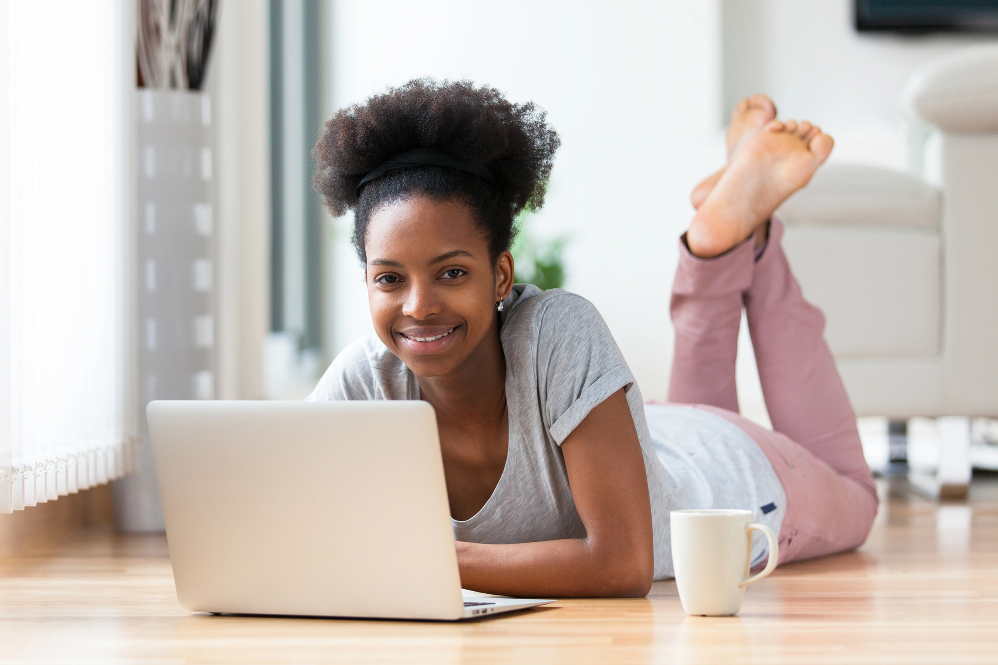 High school teen girl lying on the floor using a laptop at home with a coffee mug nearby.