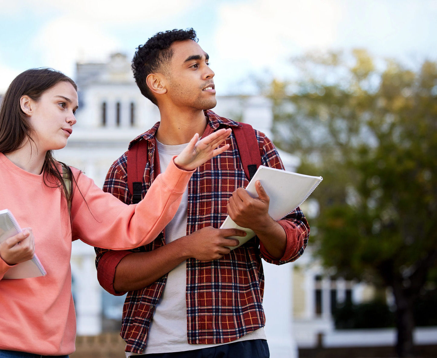 Two high school students walking on a college campus during a visit, holding notebooks and looking around as they discuss their surroundings.