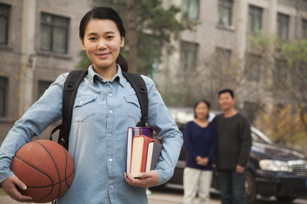 A confident high school student holding textbooks and a basketball stands in the foreground, while her supportive parents watch from a distance behind her.