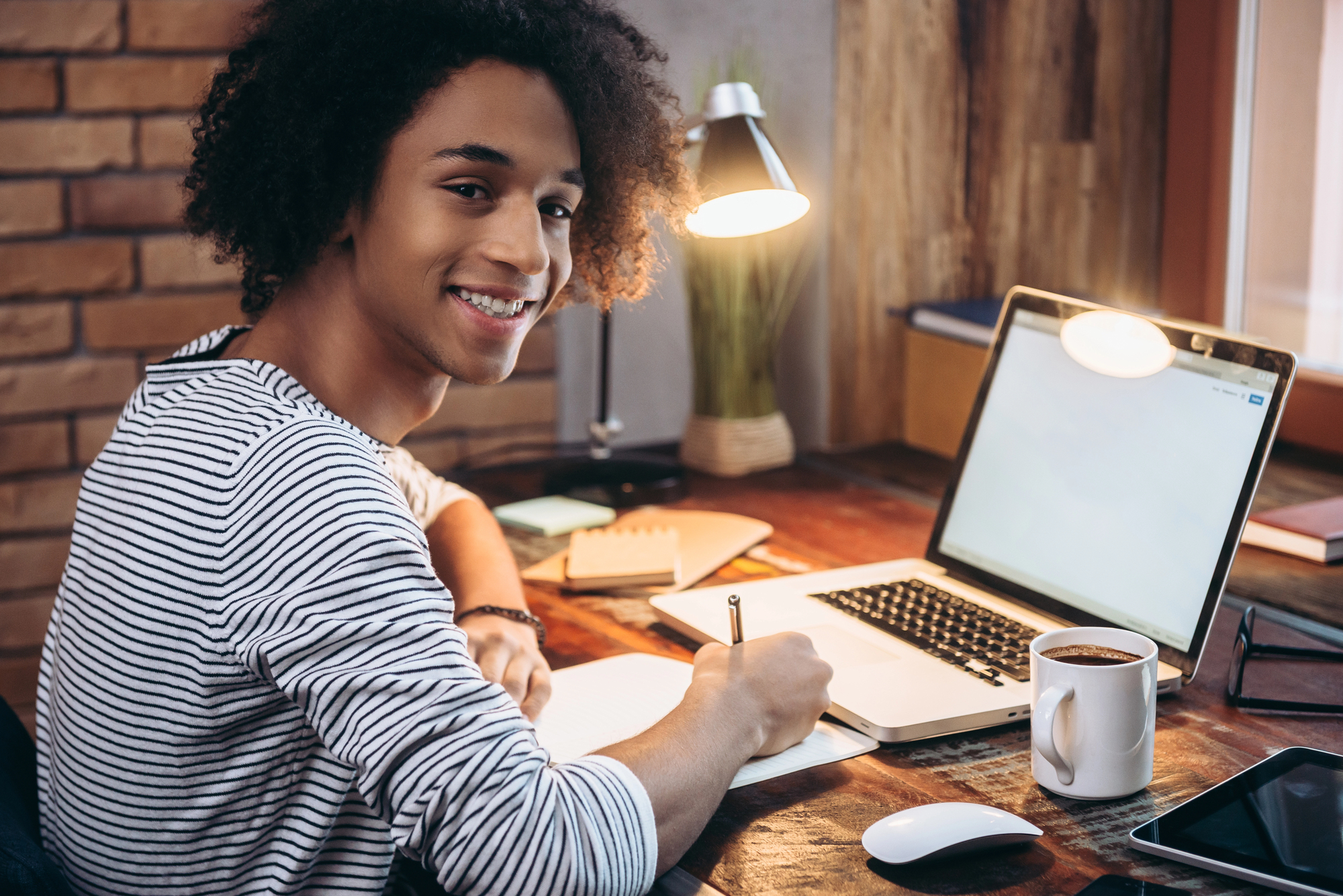Teen student smiling at a desk while writing notes beside an open laptop.