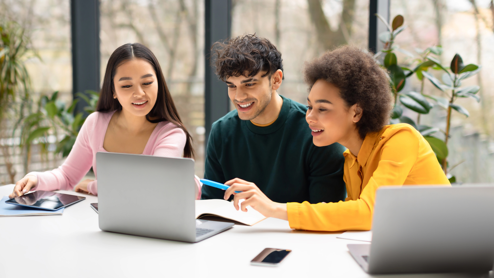 Three college students collaborate around a laptop while reviewing notes in a bright study space.